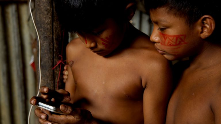 Two Indigenous children from the Amazon exploring a smartphone together indoors.